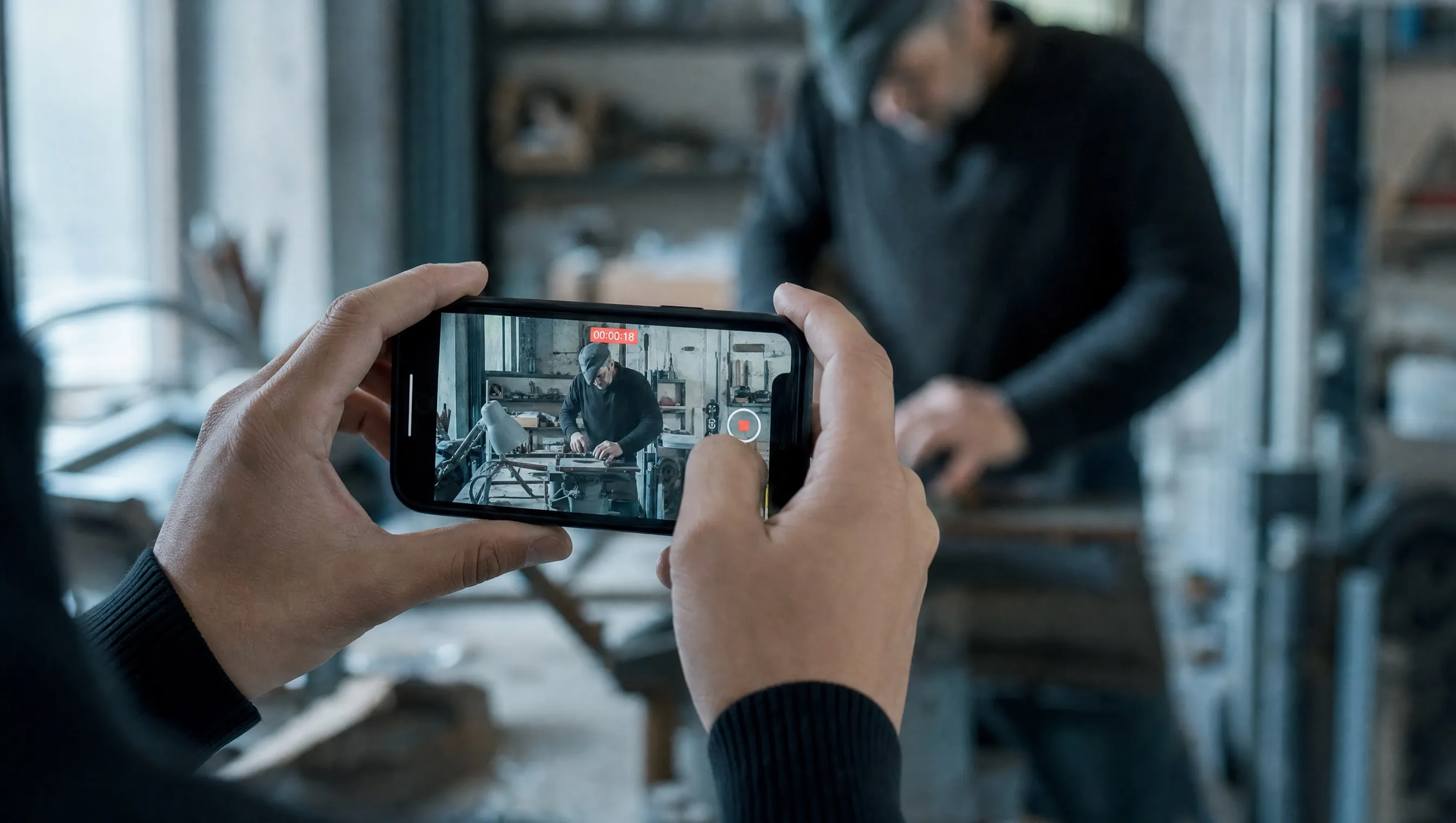 Hände filmen mit dem Smartphone einen Handwerker bei der Arbeit in einer Werkstatt
